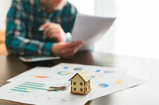 Person reviewing documents related to real estate, with a miniature house and keys on a table covered with financial charts, illustrating the role of real estate appraisers in Texas probate.