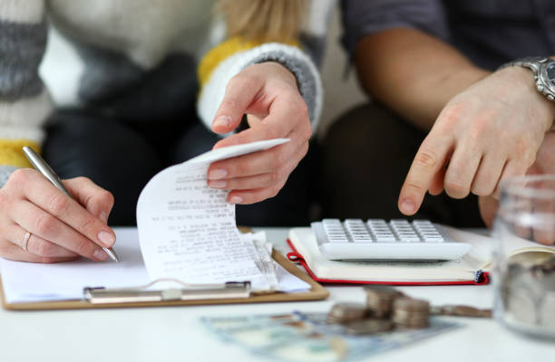 Young couple calculating finances with a calculator, reviewing expenses on a receipt, and surrounded by coins and cash, reflecting the financial challenges of dealing with probate and squatters.