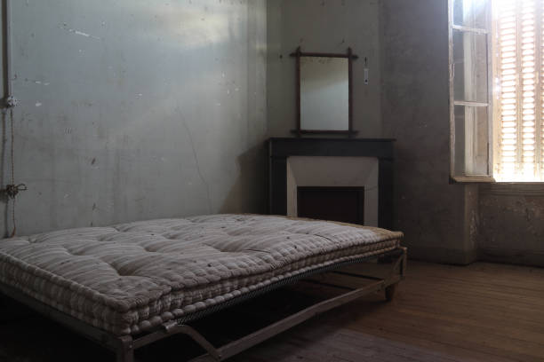 Old mattress on a metal bed frame in an empty, dimly lit room with peeling walls and a window, symbolizing the neglect and potential squatting issues in probate properties after a loved one's death.