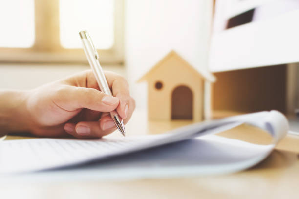 Hand signing a document related to home ownership, with a small house model in the background, symbolizing trust and estate management in Texas.