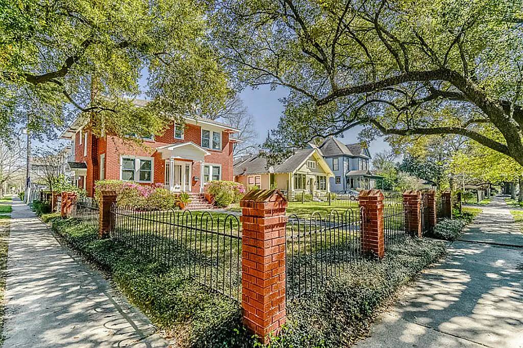 Historic residential homes along a tree-lined street in Houston, Texas, showcasing a red brick house with a white porch and vibrant landscaping, surrounded by a wrought-iron fence.